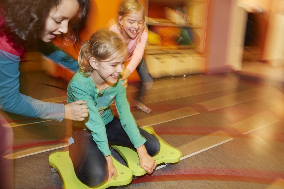 Three girls playing together: one girl sits on a rolling board while the other two push her with laughter